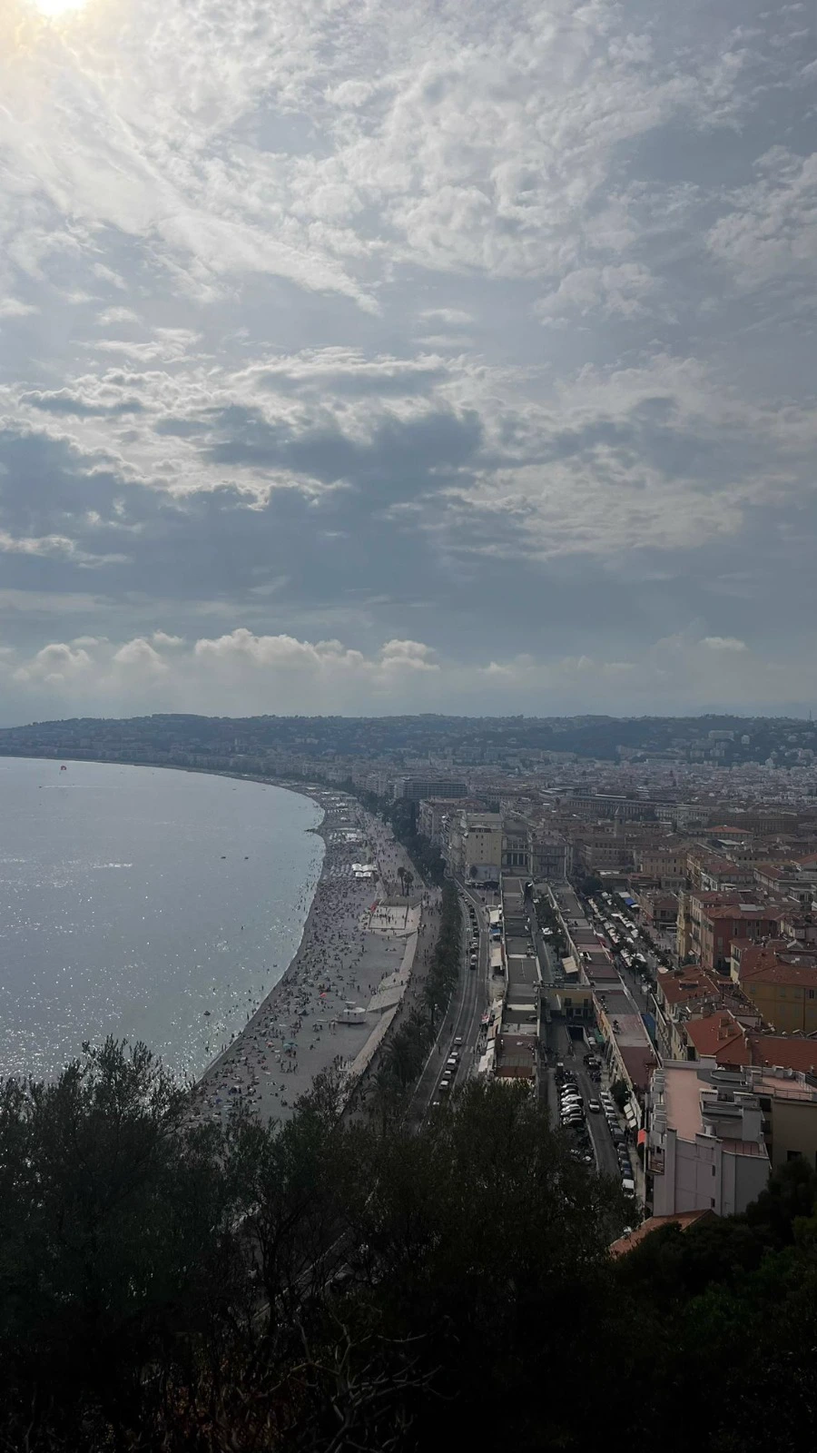 La baie de Nice vue des hauteurs, mer argentée, plage bondée et toits de tuiles sous un ciel nuageux.
