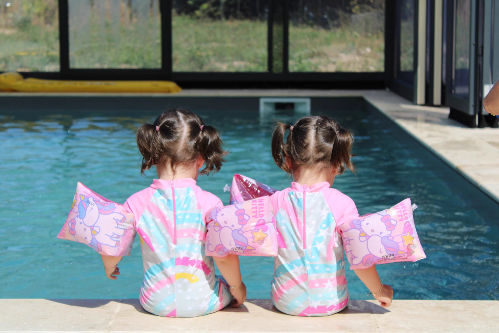 Deux petites filles de dos, assises au bord d’une piscine, brassards roses et maillots à rayures.