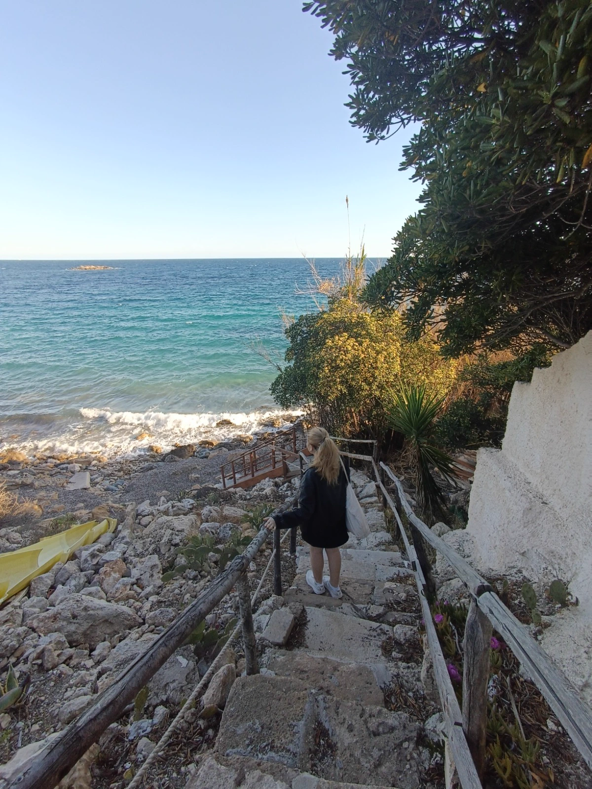 Une femme descend un escalier de pierre bordé de rambardes en bois vers une crique rocheuse et une mer turquoise.