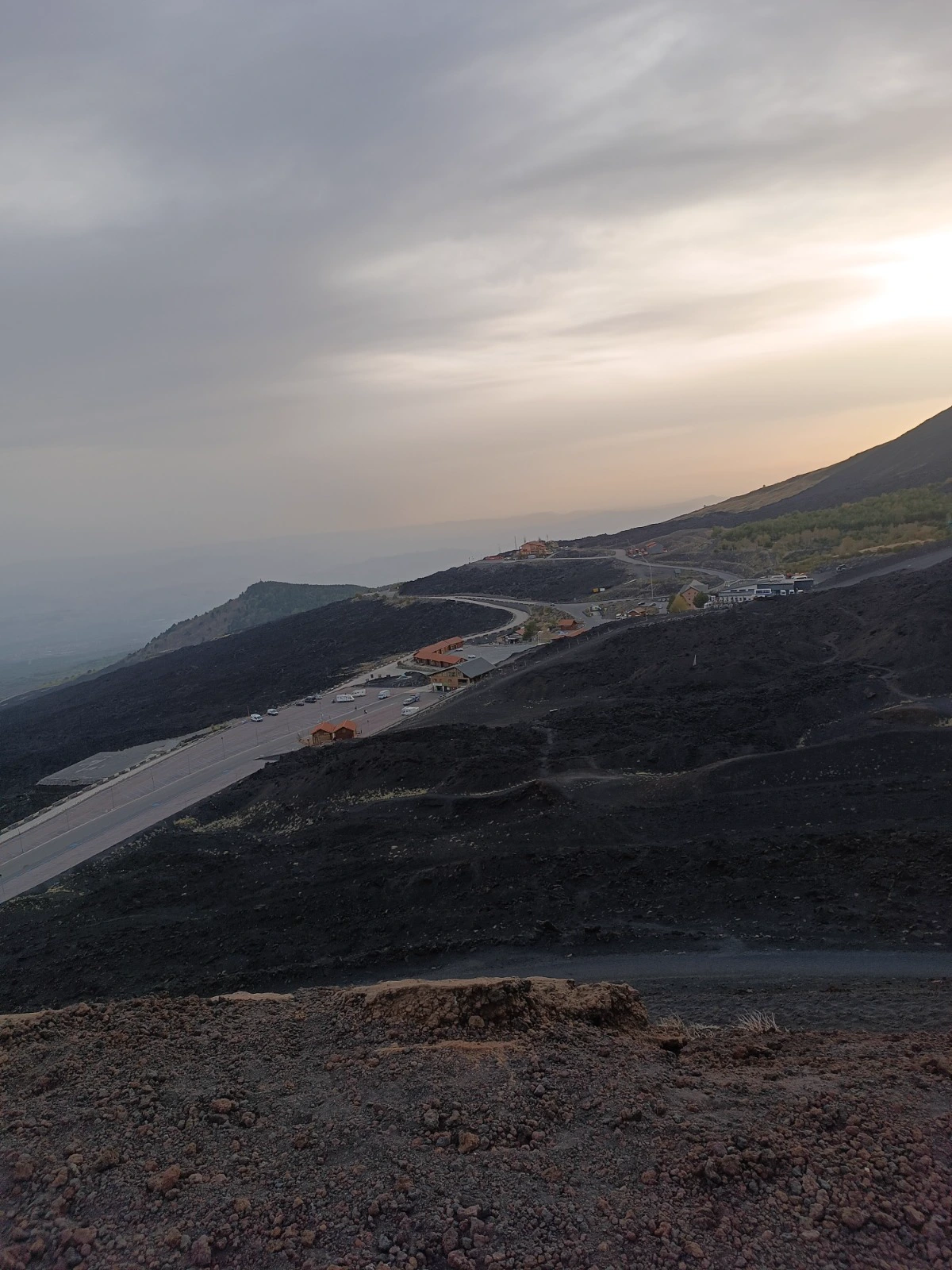 Versant de l’Etna au crépuscule, route sinueuse et refuges orange sur la lave noire, ciel rose au loin.