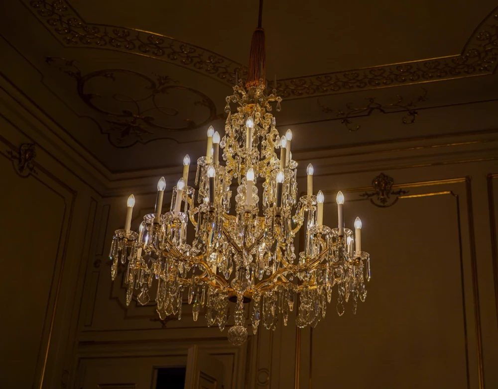 Grand lustre de cristal allumé sous un plafond à moulures dorées.