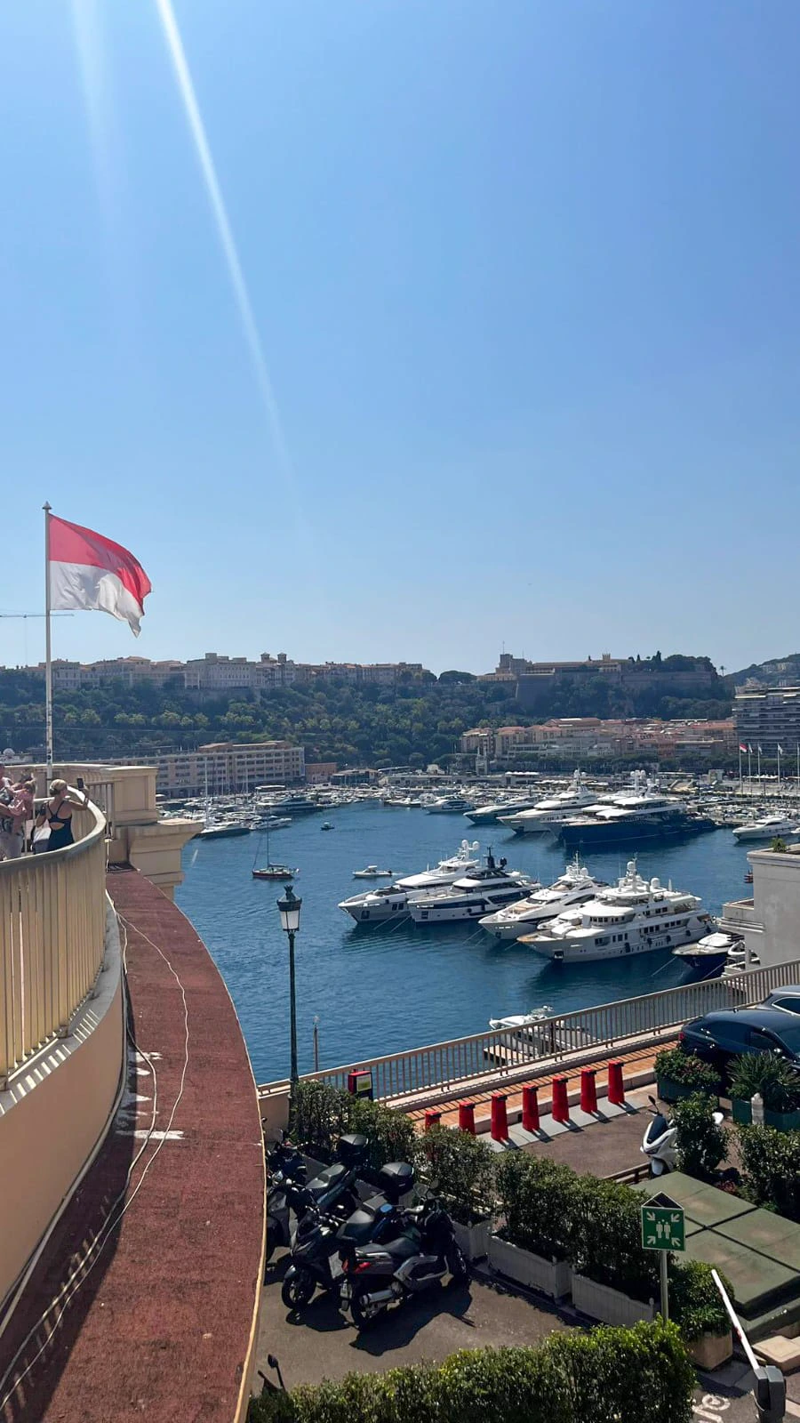 Le port de Monaco depuis une terrasse, drapeau monégasque au vent et yachts blancs alignés.