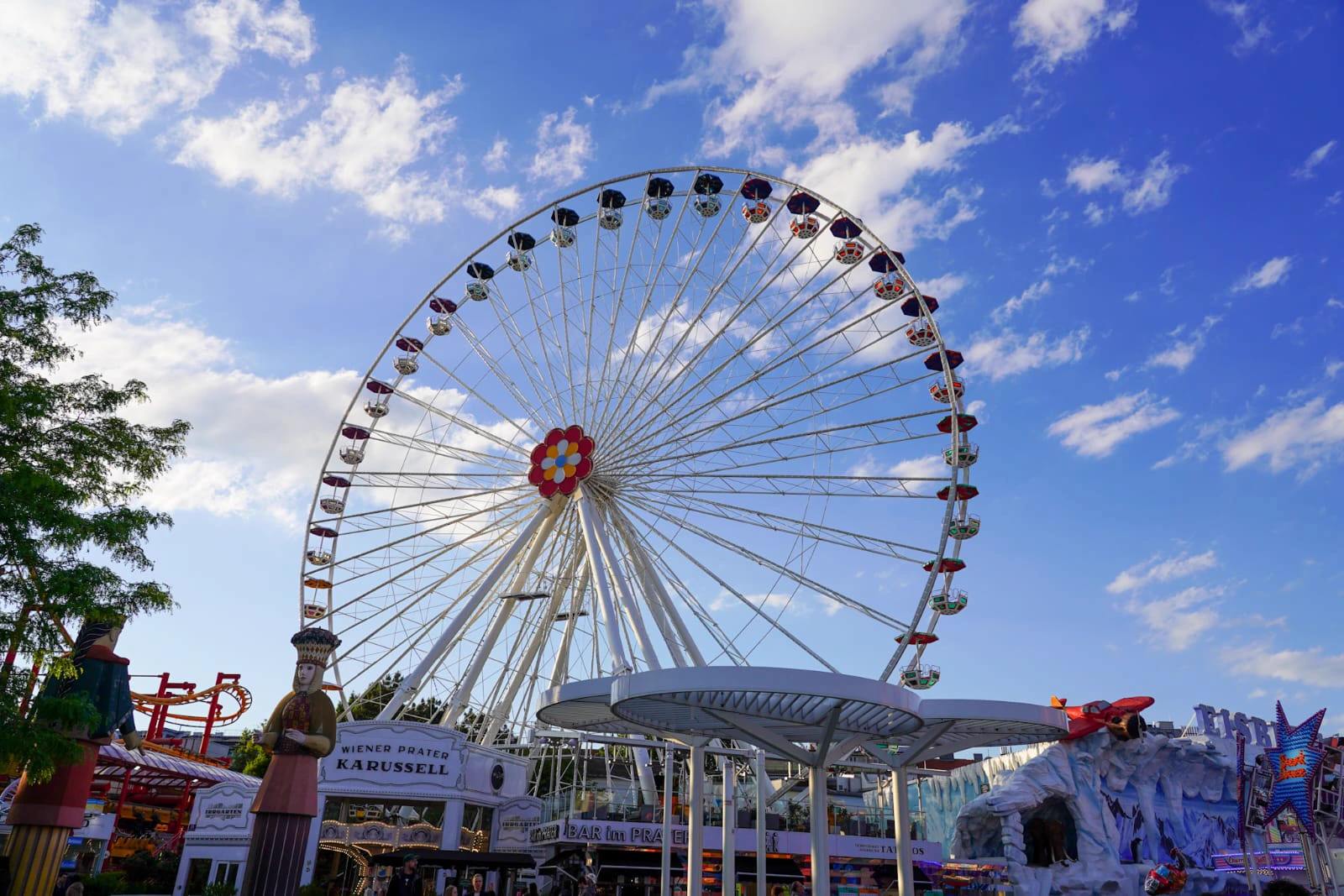 Grande roue blanche du Prater sous un ciel de nuages, enseignes de fête foraine au premier plan.